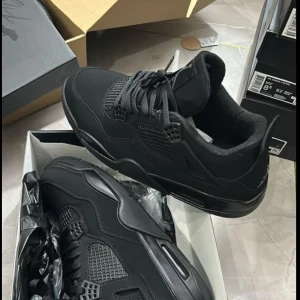 Nike Air Jordan 4 Retro Black - Säljer ett par helsvarta Nike Air Jordan 4 Retro sneakers. Skorna har en snygg och chunky siluett med meshdetaljer, snörning och ikoniska plastdetaljer på sidorna. Ovandelen är i mocka och syntet, och sulan är rejäl med synlig Air-enhet. Perfekta för dig som gillar streetwear.