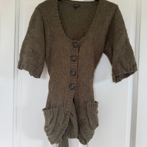 Stickad kofta från Lindex med bälte - Mysig grön stickad kofta från Lindex med korta ärmar, stora knappar framtill, två rymliga fickor och ett matchande bälte i midjan. Perfekt för lager-på-lager och chill höststil. Ullmix med akryl 