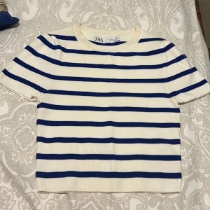 Randig ribbad topp från Zara - Vit och blå ribbad topp från Zara med korta ärmar och rund hals. Tidlös design med horisontella blå ränder på vit botten, perfekt för en fräsch och avslappnad look. Mjuk och stretchig känsla.