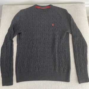 Mörkgrå kabelstickad tröja Stenströms - Mörkgrå kabelstickad tröja från Stenströms med rund halsringning och diskret röd logga broderad på bröstet. Klassisk modell med långa ärmar och ribbade muddar. Perfekt för kyliga dagar och snygg till jeans eller chinos.