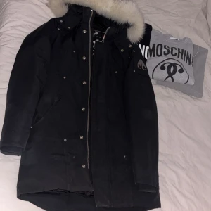 Svart parkajacka från Moose Knuckles - Svart parkajacka från Moose Knuckles med vit fuskpäls på huvan och silverfärgade knappar. Jackan har dragkedja och tryckknappar framtill, flera fickor och klassisk logga på ärmen. Perfekt för kalla dagar med sin långa modell och varma material.