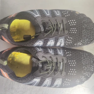 Svarta sportiga barfota sneakers med färgglad sula - Lätta och flexibla svarta barfota sneakers med ventilerande mesh, snörning och coola reflexdetaljer. Sulan är svart med färgglada inslag i orange, blått och gult. Insidan är gul och skon har en smidig passform som funkar perfekt för träning eller utomhusaktiviteter.