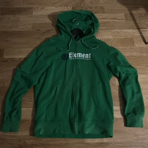 Grön hoodie från Element med tryck - Säljer en grön hoodie från Element i storlek XL. Tröjan har huva med snörning, dragkedja framtill och två fickor. På bröstet finns ett stort vitt och svart tryck med Element-loggan och texten 'wind.water.fire.earth'. Perfekt för chill dagar.