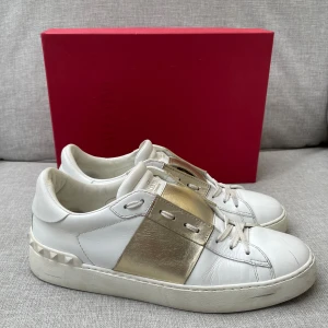 Valentino skor  - Snygga Valentino Garavani Open sneakers i vitt skinn med metallicguld detalj över snörningen. Klassisk rund tå, platt sula och ikoniska nitar baktill. Perfekta för dig som vill sticka ut med en lyxig touch i vardagen.