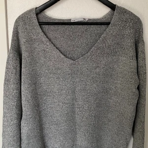 Grå stickad tröja från 157 - Mysig grå stickad tröja från 157 med v-ringad hals och lång ärm. Perfekt för lager-på-lager och chill dagar. Tröjan har en enkel och stilren design som funkar till det mesta. Säljes i storlek S.