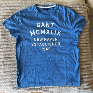 Blå t-shirt från Gant med tryck - Snygg blå t-shirt från Gant med vit texttryck på bröstet: 'GANT MCMXLIX NEW HAVEN ESTABLISHED 1949'. Klassisk rund halsringning och korta ärmar. Perfekt för en avslappnad och stilren look.