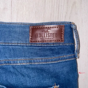 Blå high-rise jeans från Hollister - Snygga blå high-rise jeans från Hollister med slitna detaljer och hål på ena benet. Modellen är super skinny och har klassiska bakfickor samt gylf med knapp. Perfekta för en avslappnad och trendig look.