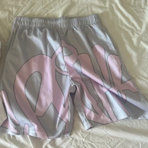 Grå syns world - Säljer ett par grå shorts med bred resår i midjan och ett stort, rosa grafiskt tryck på framsidan. Shortsen har en loose passform och är tillverkade i ett syntetmaterial som känns lätt och luftigt. Perfekta för sommaren när du vill sticka ut lite extra.
