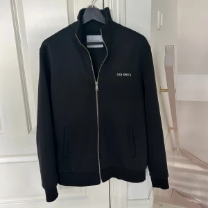 Svart zip tröja från Les Deux - Säljer en svart zip tröja från Les Deux med stilren design och vit broderad logga på bröstet. Tröjan har dragkedja framtill, ståkrage och två fickor. Perfekt för dig som gillar en clean och sportig look. Detta är den gamla designen utan summar i mitten av tröjan, som inte görs längre.