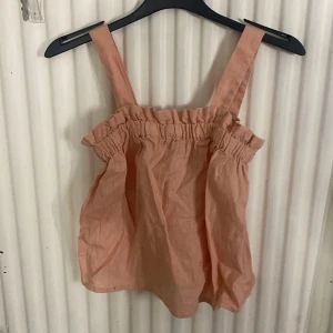 Rosa linnetopp med volangkant - Söt rosa linnetopp med breda axelband och volangkant upptill. Toppen har en luftig och loose passform, perfekt för varma dagar. Materialet är linne och färgen är en mjuk, ljusrosa ton. Enkel och trendig design som ger en chill vibe.