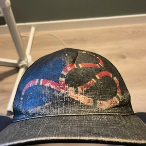 Svart keps från Gucci med ormtryck - Svart keps från Gucci med ikoniskt röd och beige ormtryck framtill. Baksidan har justerbart band med röd och svart randig detalj. Kepsen är tillverkad i mesh och syntetmaterial för en luftig känsla. Perfekt accessoar för att lyfta din streetwear-stil.