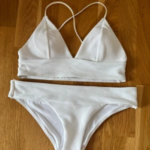 Vit ribbad bikini med trekantsöverdel - Säljer en stilren vit bikini med ribbad struktur och guldiga detaljer. Överdelen har trekantskupor och smala axelband som korsas i ryggen. Underdelen är klassisk med bredare kant upptill. Perfekt för stranden eller poolen på sommarn eller semester eller varför inte nu i kylan i en go yacutsy eller bastu. Passar str 34-38