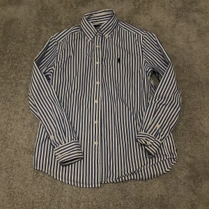 Randig skjorta från Ralph Lauren - Snygg skjorta från Ralph Lauren med blå och vita vertikala ränder, klassisk krage och knappar framtill. Skjortan har lång ärm och ikonisk broderad logga på bröstet. Tillverkad i mjuk bomull som känns skön mot huden.