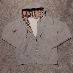 Grå hoodie från Burberry med dragkedja - Säljer en grå hoodie från Burberry med klassiskt rutigt foder i huvan och insidan. Tröjan har dragkedja, snörning i huvan och broderad logga på bröstet. Perfekt för dig som gillar stilrena och lyxiga streetwear-plagg.