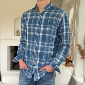  Denim goods Polo Ralph Lauren skjorta - Snygg blå och vit rutig skjorta från Polo Ralph Lauren med klassisk button-down krage och broderad logga på bröstet. Skjortan är i bomull och har långa ärmar samt knäppning framtill. Perfekt för en chill och stilren look.