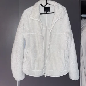 Vit teddyjacka från 157 - Fluffig vit teddyjacka från 157 i storlek M. Jackan har en mjuk och gosig yta, hög krage och hel dragkedja framtill. Två sidofickor och långärmad modell. Perfekt för dig som gillar chill och comfy stil.använd 2 gånger och säljer för att den är för stor för mig