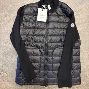 Moncler Cardigan  - Svart dunjacka från Moncler med quiltad front och stickade ärmar. Jackan har dragkedja, ribbad krage och Moncler-logga på ärmen. Insidan har ett coolt serietryck och flera etiketter. Perfekt för dig som vill ha en snygg och bekväm jacka med street vibe.