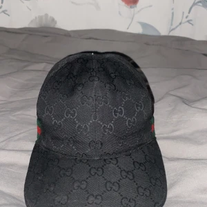 Svart Gucci keps med GG-mönster - Snygg svart keps från Gucci med det klassiska GG-monogrammet över hela kepsen. Kepsen har justerbar rem baktill i svart skinn och diskreta gröna och röda detaljer på sidorna. Materialet är bomull och kepsen har en rundad skärm för en clean look. PS - Knoppen fattas.