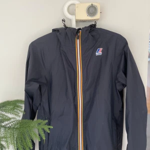 Svart vindjacka från K-Way med huva - Svart vindjacka från K-Way med huva och justerbara snören. Jackan har en färgglad dragkedja med ränder i gult, orange och vitt samt K-Way-logga på bröstet. Lätt och smidig design, perfekt för blåsiga dagar. Långärmad och tillverkad i syntetmaterial.