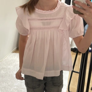 Ljusrosa blus med volang från Gina Tricot - Supersöt ljusrosa blus från Gina Tricot med volangdetaljer på axlarna och runt halsen. Blusen har korta puffärmar, dekorativa broderade band framtill och knäppning bak i nacken. Perfekt för en söt och feminin look.