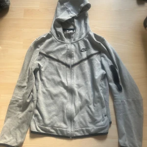 Grå Nike hoodie med dragkedja XL 158-170cm - Snygg grå hoodie från Nike i storlek XL med hel dragkedja och huva. Tröjan har svarta detaljer längs bröstet och ärmarna samt diskret Nike-logga på bröstet. Perfekt för chill eller träning, med bekväm passform och praktiska fickor. Pris kan diskuteras.