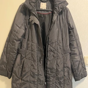 Grå quiltad dunjacka från Cappuccini - Snygg grå dunjacka från Cappuccini med quiltad design och huva. Jackan har dragkedja framtill, praktiska fickor och markerad midja för en schysst siluett. Perfekt för kalla dagar när du vill hålla stilen och värmen.