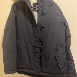 Mörkblå parkajacka från Race Marine - Parkajacka i mörkblått från Race Marine med huva och snygg fuskpäls runt kragen. Jackan har stora knappar framtill, dragkedja och två fickor med lock. Badge på ena ärmen och fodrad insida. Perfekt för kalla dagar.