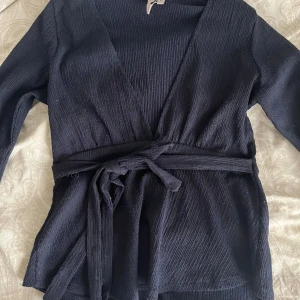 Mörkblå Nelly blus  - Säljer en svart omlottopp från Nelly i storlek XS. Toppen har lång ärm, knytband i midjan och en djup v-ringning. Materialet är strukturerat och ger en snygg textur. Perfekt för dig som gillar stilrena och trendiga plagg.