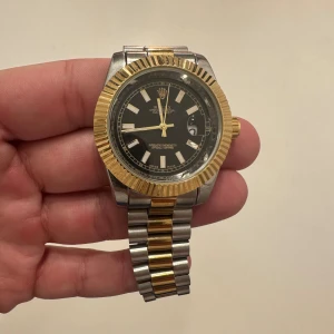 Tvåtonad klocka - Snygg klocka med svart urtavla, guldfärgade detaljer och tvåtonad länk i silver och guld. Klockan har klassisk räfflad bezel i guld och tydliga indexmarkeringar. Länken är i rostfritt stål och guld, med logga på spännet. Perfekt för dig som gillar exklusiv stil.