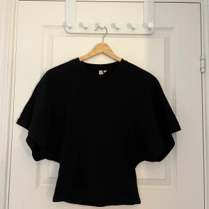 Svart topp med vida ärmar - En svart topp med rund halsringning och extra vida, korta ärmar. T-shirten har en rak passform och är tillverkad i ett mjukt material som känns skönt mot huden. Perfekt för en avslappnad och stilren look. Endast testad 