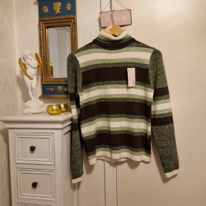 Randig polotröja från Jiangqianqian stickad tröja  - Snygg polotröja från Jiangqianqian med breda ränder i grönt, brunt och vitt. Tröjan har ribbad polokrage och långa ärmar. Materialet är mjukt och stickat, perfekt för dig som gillar retrovibbar och coola färgkombinationer. Köpt I Italien.