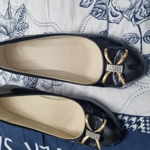 Svarta lackpumps med rosett och klack - Snygga svarta pumps i lack med rund tå och låg klack. Framtill sitter en dekorativ rosett i svart och guld med glittrande stenar. Skorna har en elegant och klassisk look, perfekta för att lyfta din outfit.
