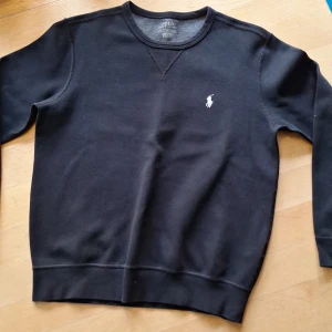 Svart sweatshirt från Polo Ralph Lauren - Svart sweatshirt från Polo Ralph Lauren i bomull med klassisk rund hals och vit broderad logga på bröstet. Tröjan har långa ärmar och ribbade muddar vid ärmslut och nederkant. Perfekt för en chill och stilren look.