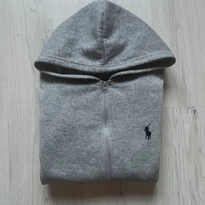 Ralph lauren zip  - Snygg grå hoodie från Polo Ralph Lauren med dragkedja och huva. Klassisk svart logga på bröstet och två fickor framtill. Perfekt för chill dagar och enkel att matcha med jeans eller joggers. Mjuk bomullskvalitet och ribbade muddar vid ärmslut och nederkant.