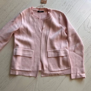 Ljusrosa cardigan jacka från Gina Tricot - Superfin ljusrosa cardigan jacka från Gina Tricot i stickat material. Jackan har raka linjer, två stora fickor framtill och en öppen design utan knappar eller dragkedja. Perfekt för lager-på-lager och ger en clean look.