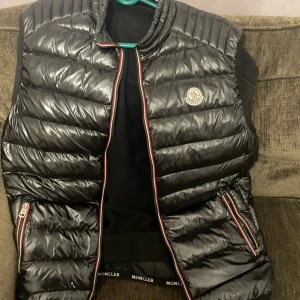 Svart dunväst från Moncler , M/L - Snygg svart dunväst från Moncler med quiltad design och glansig finish. Västen har dragkedja framtill och två sidofickor med dragkedja, samt Moncler-logga på bröstet. Rödvit kant längs dragkedjan ger en sportig touch.  Obs. Dragkedjan är sönder men går att fixa hos en skräddare . 