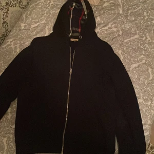Svart Burberry tröja storlek M passar i S - Svart hoodiejacka från Burberry med klassiskt rutigt foder i huvan. Jackan har dragkedja framtill, broderad logga på bröstet och en clean, stilren look. Perfekt för dig som gillar exklusiva streetwear-vibbar och snygga detaljer.