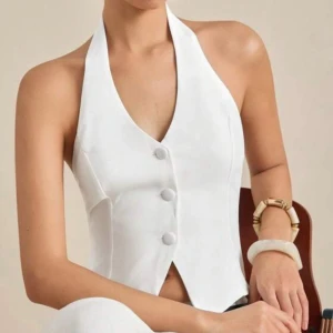 Vit omlottväst med knyt och knappar - Snygg vit väst med halterneck och v-ringad front. Västen har tre stora knappar framtill och en öppen rygg med knytband som ger en cool omlott-look. Passformen är croppad och materialet känns slätt och lätt, perfekt för varmare dagar. Har aldrig använt den så den är så gott som ny, dock utan prislapp. Storlek S kan även funka en xs 