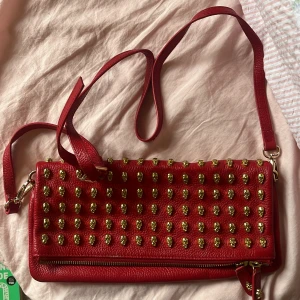 Röd axelväska med gulddetaljer - Säljer en röd axelväska från Alexander McQueen i skinn med coola guldfärgade dödskalle-nitar på framsidan. Väskan har dragkedja upptill och ett mönstrat innerfoder med logga. Perfekt storlek för det viktigaste och riktigt edgy vibe.