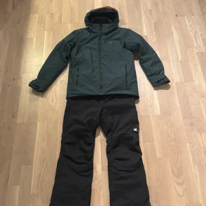 Mörkgrön Helly Hansen vinterjacka samt skidbyxor - Säljer en mörkgrön vinterjacka från Helly Hansen med huva och hel dragkedja framtill. Jackan har två sidofickor med dragkedja och justerbara ärmslut. Perfekt för kalla dagar och outdoor-aktiviteter. Finns möjlighet att även köpa de svarta skidbyxorna från Everest