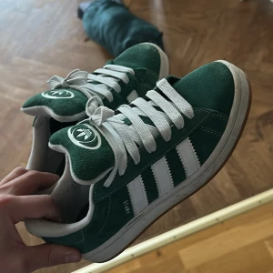 Adidas Stan Smith gröna sneakers - Gröna Adidas Stan Smith sneakers i mocka med klassiska vita ränder och vit sula. Skorna har rund tå, platt sula och vita snören. Ikonisk Adidas-logga på plösen och snygg retrostil som passar perfekt till streetwear.
