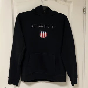 Svart hoodie från GANT med logga - Svart hoodie från GANT med broderad logga och sköld på bröstet. Klassisk känguruficka framtill och huva. Perfekt för dig som gillar stilren streetwear med preppy touch.
