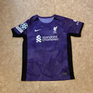 Liverpool FC Salah #11 lila matchtröja - Liverpool FC fotbollströja i lila med svarta detaljer, tillverkad av lätt och ventilerande material. Tryck med Standard Chartered, Champions League-märke på ärmen och M. Salah #11 på ryggen. Snyggt mönster och klassisk Nike-logga på bröstet.