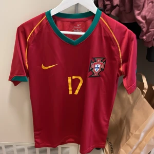 Portugal C. Ronaldo #17 Nike fotbollströja - Snygg Portugal fotbollströja från Nike med C. Ronaldo och nummer 17 på ryggen. Tröjan är röd med gula detaljer, gröna inslag vid krage och ärmslut samt Portugals landslagsemblem på bröstet. Tillverkad i lätt och ventilerande polyester, perfekt för match eller träning.