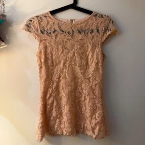 Ljusrosa topp Chiara Forthi - Säljer en ljusrosa topp från Chiara Forthi Milano. Toppen har ett vackert blommigt spetsmönster, korta ärmar och en snygg dragkedja i ryggen. Detaljerad med en liten guldfärgad metallbricka på ärmen. Perfekt för dig som gillar romantisk stil.