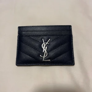 Svart YSL plånbok  - Snygg svart plånbok från YSL med silverfärgad logga framtill. Plånboken har quiltad design och flera kortfack, perfekt för att hålla koll på dina kort och cash. Stilren och lyxig accessoar som lyfter din look. Pris går att  diskutera vid snabba affärer då den inte kommer till användning 