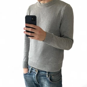 Grå stickad tröja från Dobber | M - Snygg grå stickad tröja från Dobber i storlek M. Tröjan har rund hals, ribbade muddar vid ärmslut och nederkant samt långärmad passform. Perfekt för dig som gillar enkel och stilren look med mjukt material.
