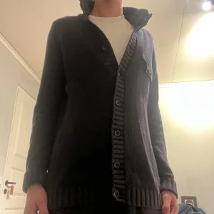 Mörkblå stickad kofta med knappar - Säljer en mörkblå stickad kofta med ribbade detaljer och stora knappar framtill. Koftan har lång ärm och en liten bröstficka med knapp. Perfekt för dig som gillar chill och stilren look.