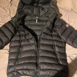 Svart dunjacka från Colmar med huva - Snygg svart dunjacka från Colmar med huva och quiltad design. Jackan har dragkedja framtill, två sidofickor med dragkedja och en innerficka. Perfekt för kalla dagar och har en clean, sportig look. Finns ett litet hål ovanför axeln syns knappast när man har jackan på! Vid snabb affär kan priset diskuteras
