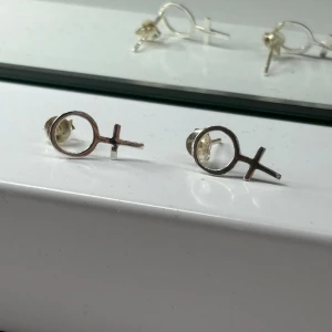 Silvriga örhängen med venus-symbol, kvinna - Säljer ett par stilrena örhängen i silverfärg formade som venus-symbolen. De är små och minimalistiska, perfekta för dig som vill ha ett statement men ändå hålla det enkelt. Stiftmodell som sitter bekvämt i örat.